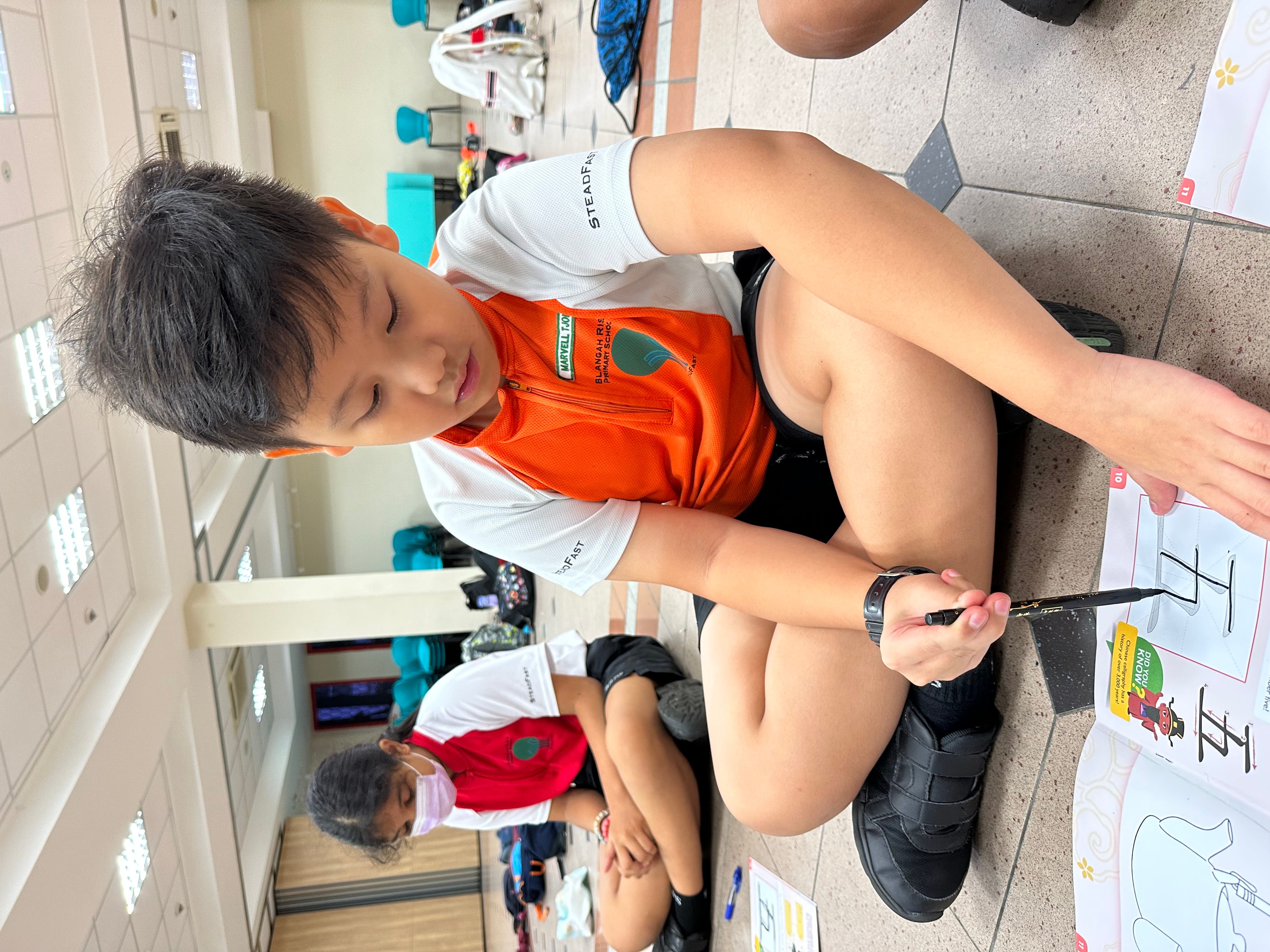 Boy in orange shirt sits on tiled floor drawing with a brush on paper; masked child sits behind.